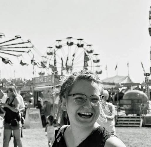 Nat laughs at something just off camera in a black and white photo. In the background there is a fair in full swing with people walking about, a Ferris wheel and giant swing all caught mid movement.